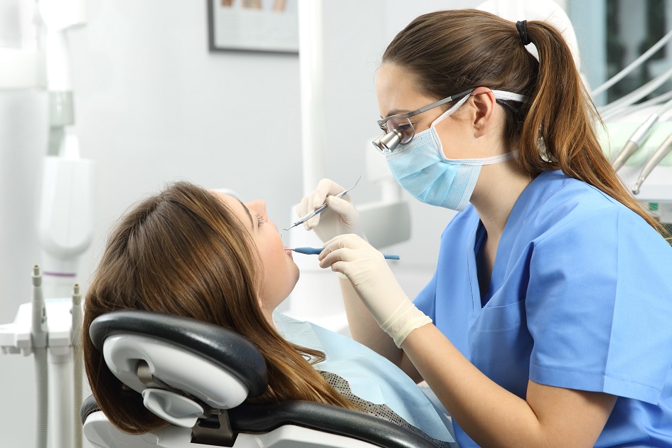Dentista vestindo óculos, máscara e jaleco azul está examinando a paciente que está sentada na cadeira. Ambas estão no consultório do dentista.